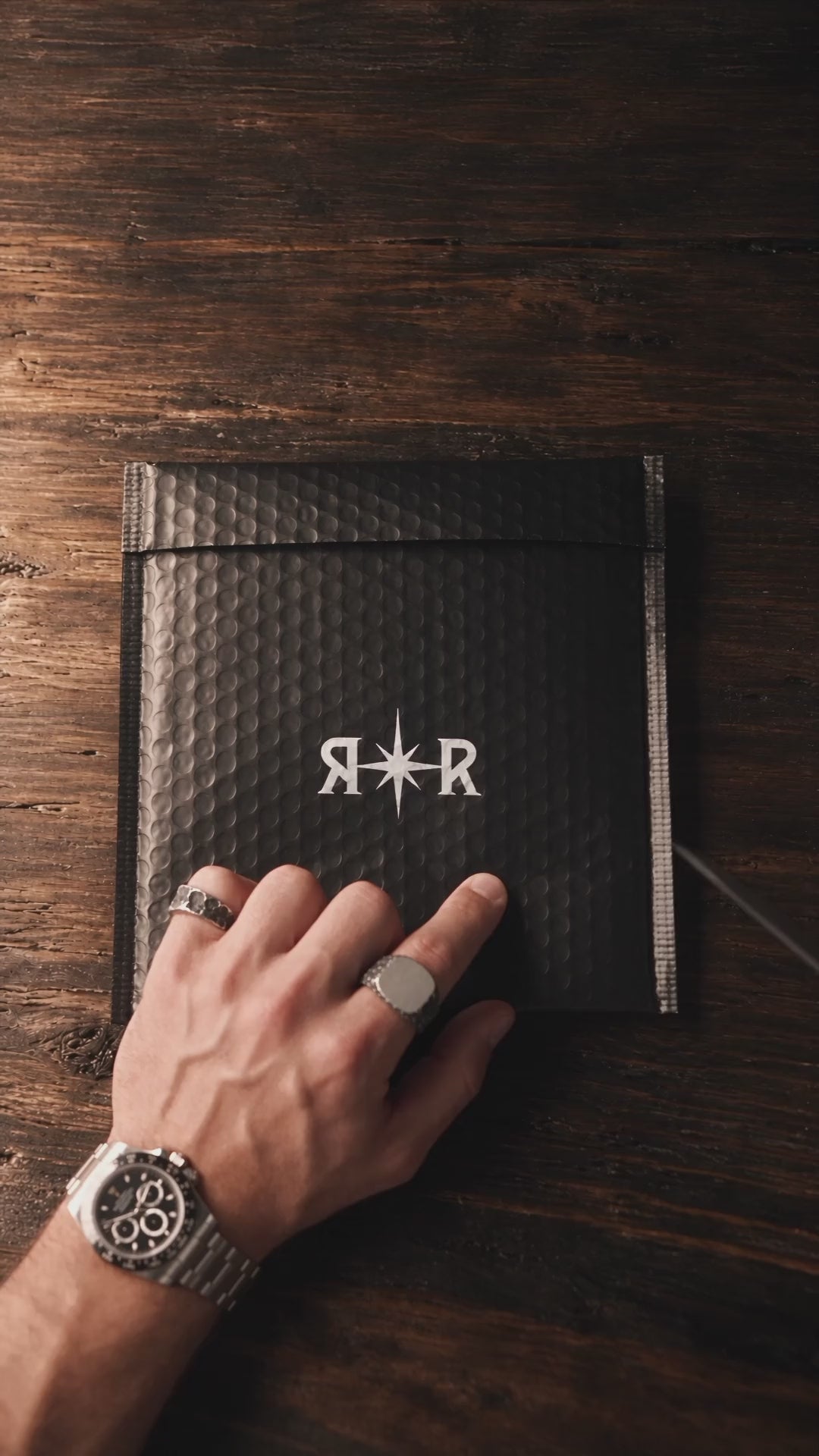 Unboxing from above on a walnut table: the black R✦R mailer, the wax-sealed envelope with a letter and an aluminium card, the ring box drawn out of its sleeve, and the Raw I Ring going onto a tattooed hand.