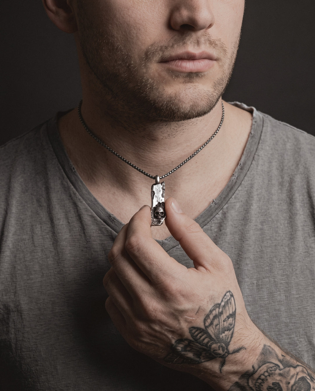 Raw I Pendant worn by a man, held between his thumb and index finger, a moth tattoo on the back of his hand