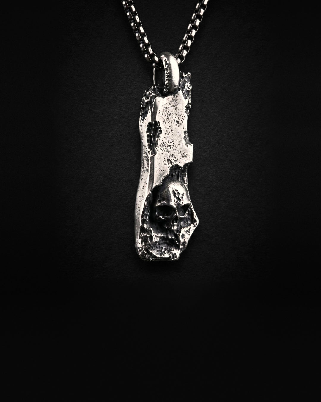 Raw I Pendant at an angle, the pitted edges of the bar and the skull at its foot, on a box chain