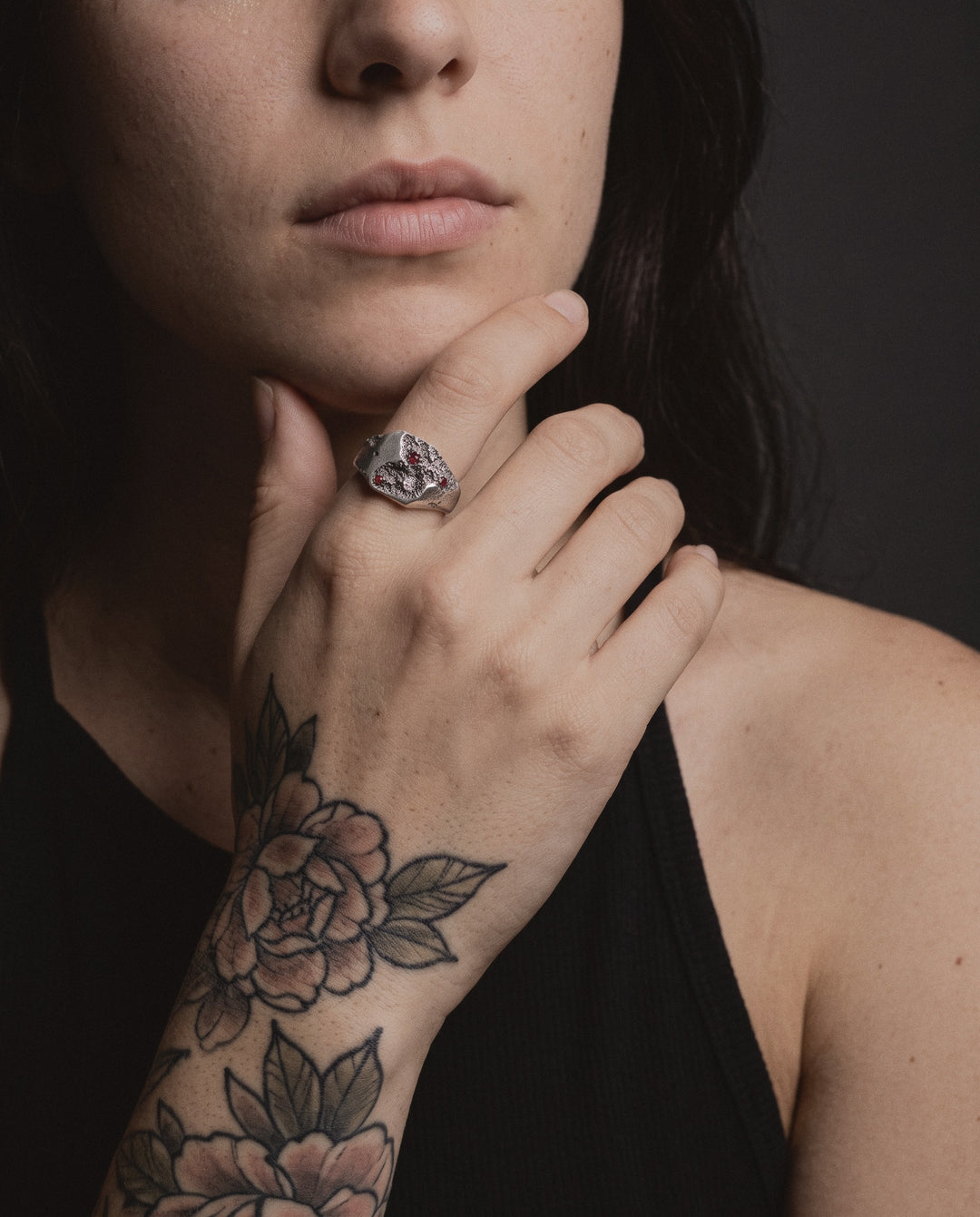 Raw I Ring with ruby stones worn by a woman on her index finger, her hand at her chin, a neo-traditional peony tattoo on the back of her hand
