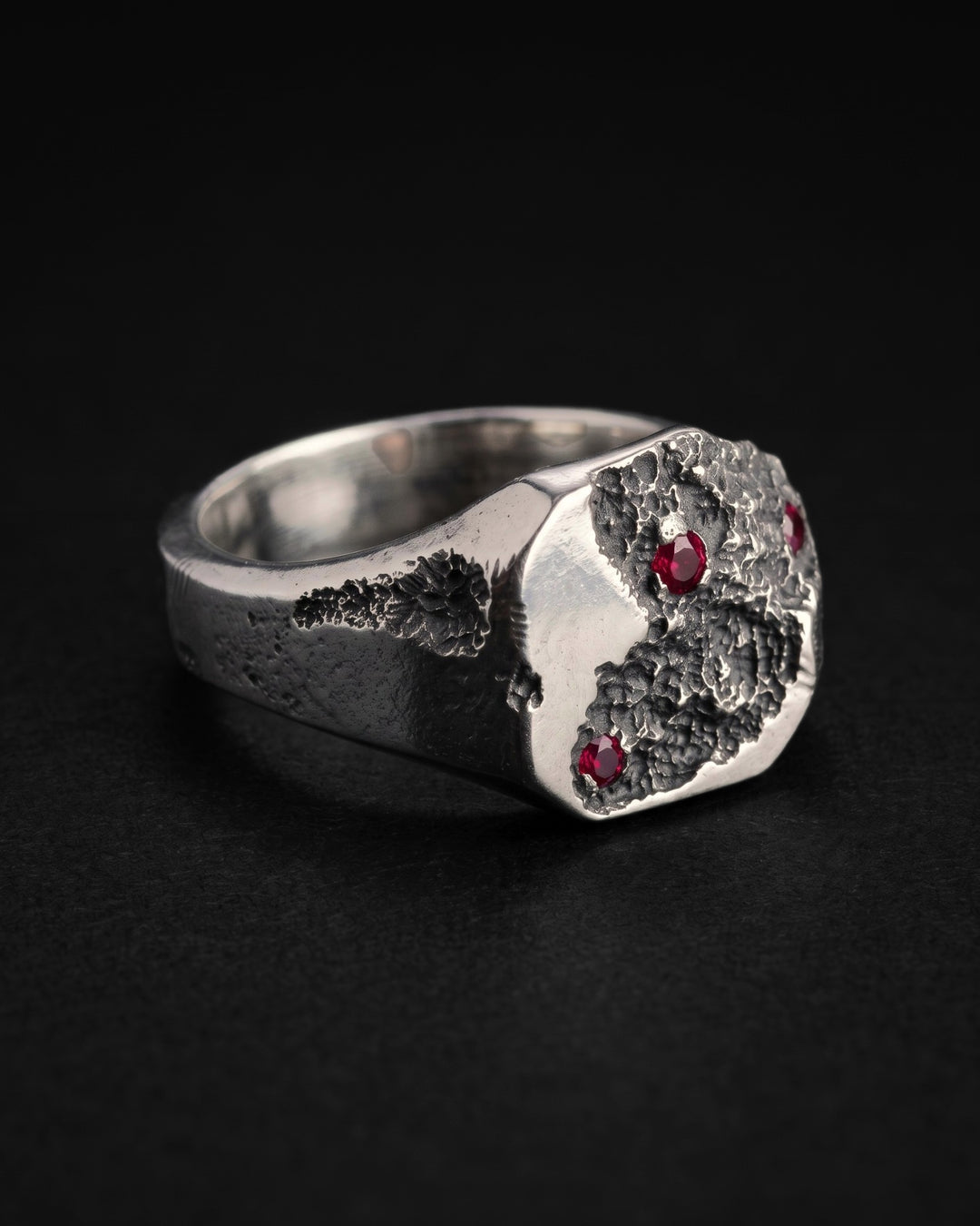 Raw I Ring with ruby stones at an angle on black, the cratered signet face with its three rubies and the mirror polished inside