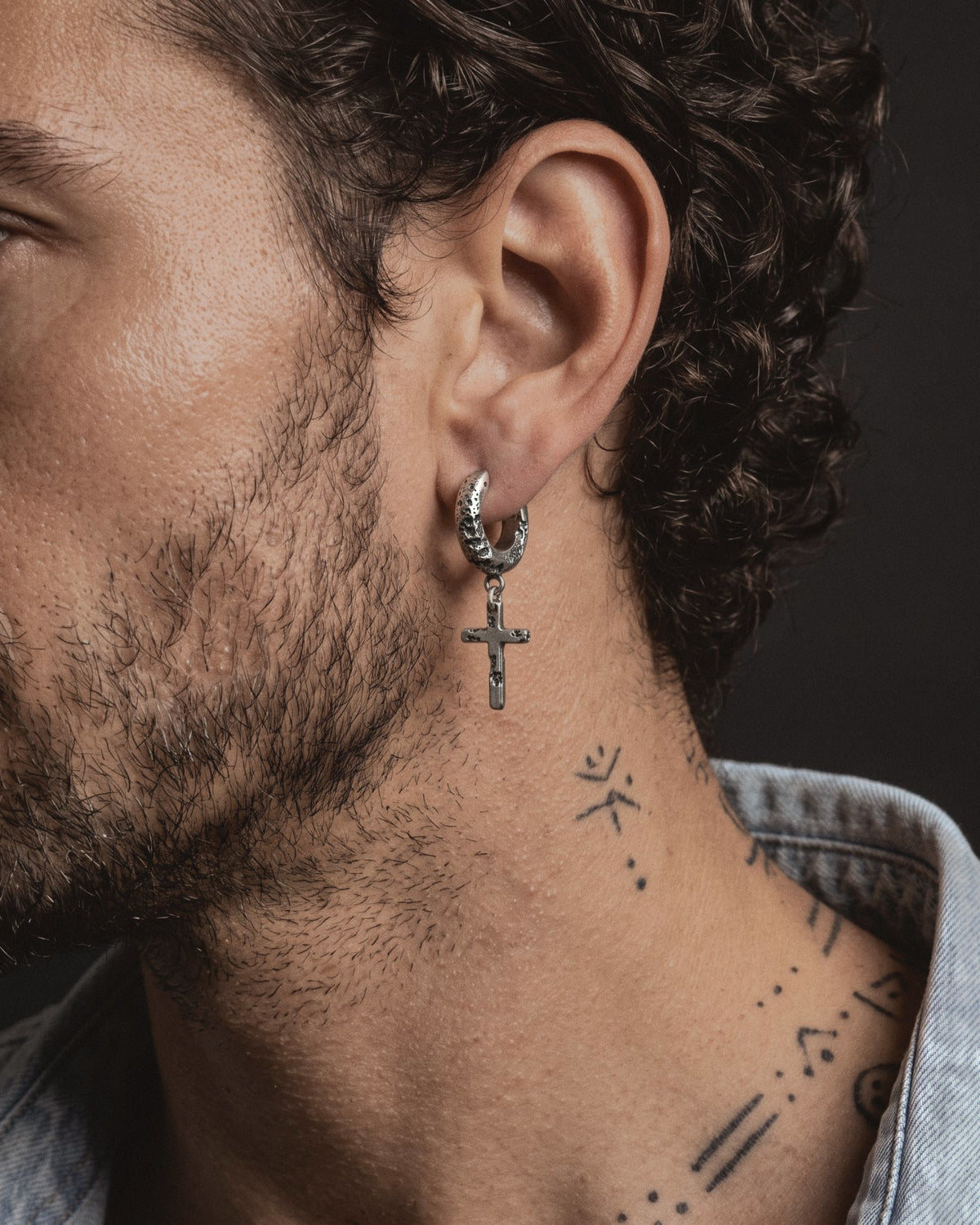 Raw II Earring worn by a man, the small cratered cross hanging below the silver hoop, with a denim shirt and fine neck tattoos