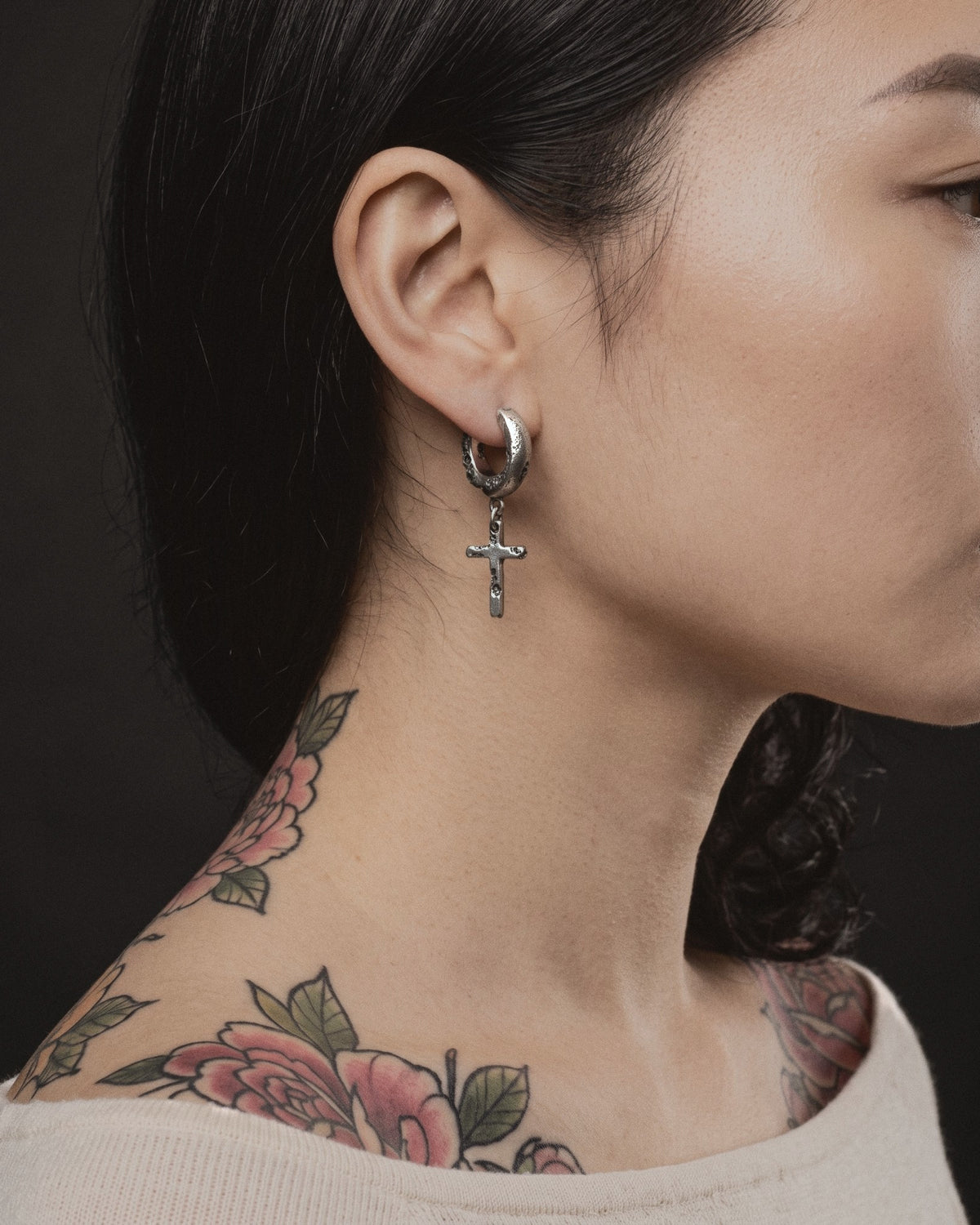 Raw II Earring worn by a woman, the cratered silver hoop and small cross above colour peony tattoos on her neck and shoulder