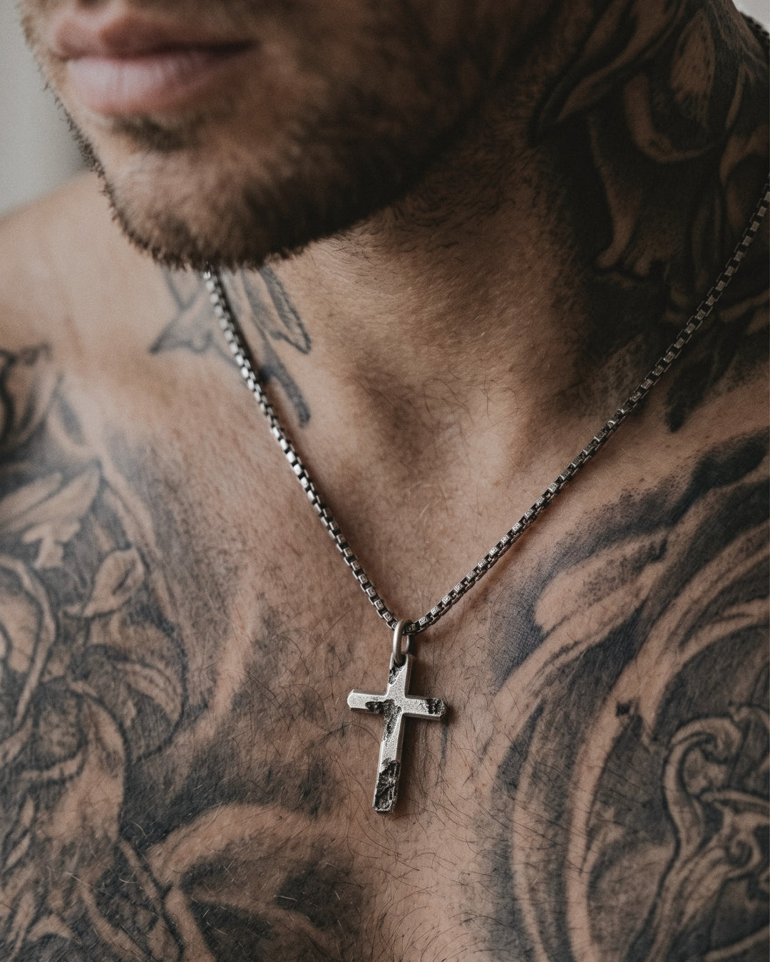 Raw II Pendant, the small cratered silver cross on a box chain, worn on a man's tattooed chest