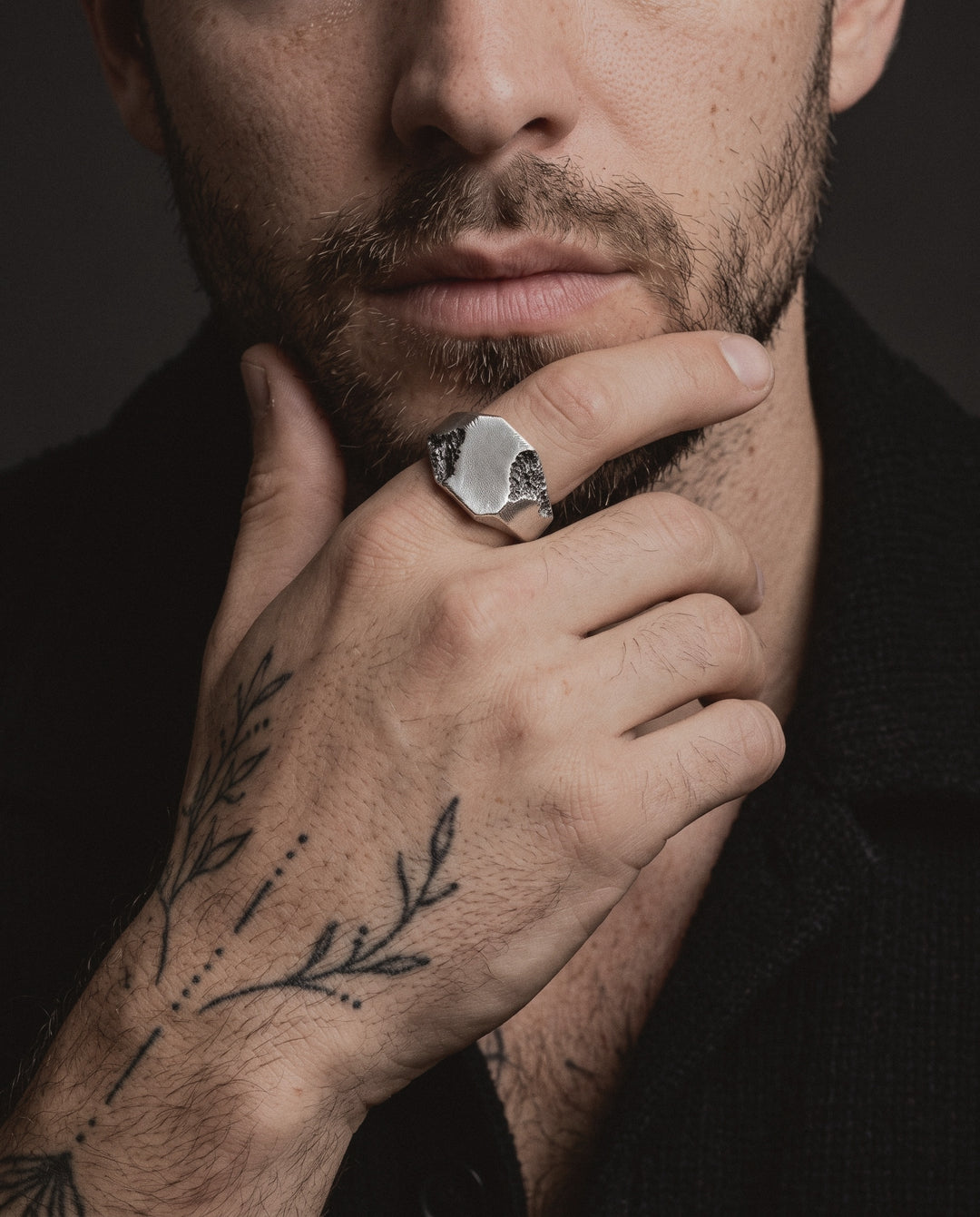 Raw II Ring worn by a man on his index finger, his hand at his chin, fine line botanical tattoos on his hand and forearm