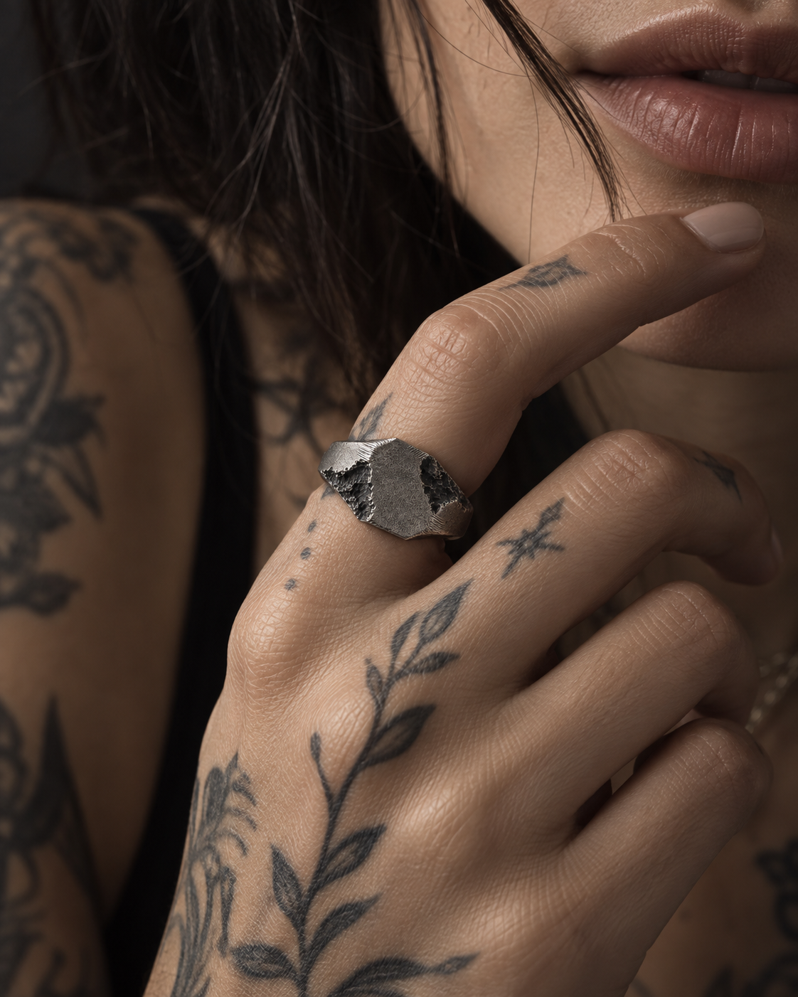 Raw II Ring on a woman's tattooed hand, signet plate turned to the camera, hand resting at her chin