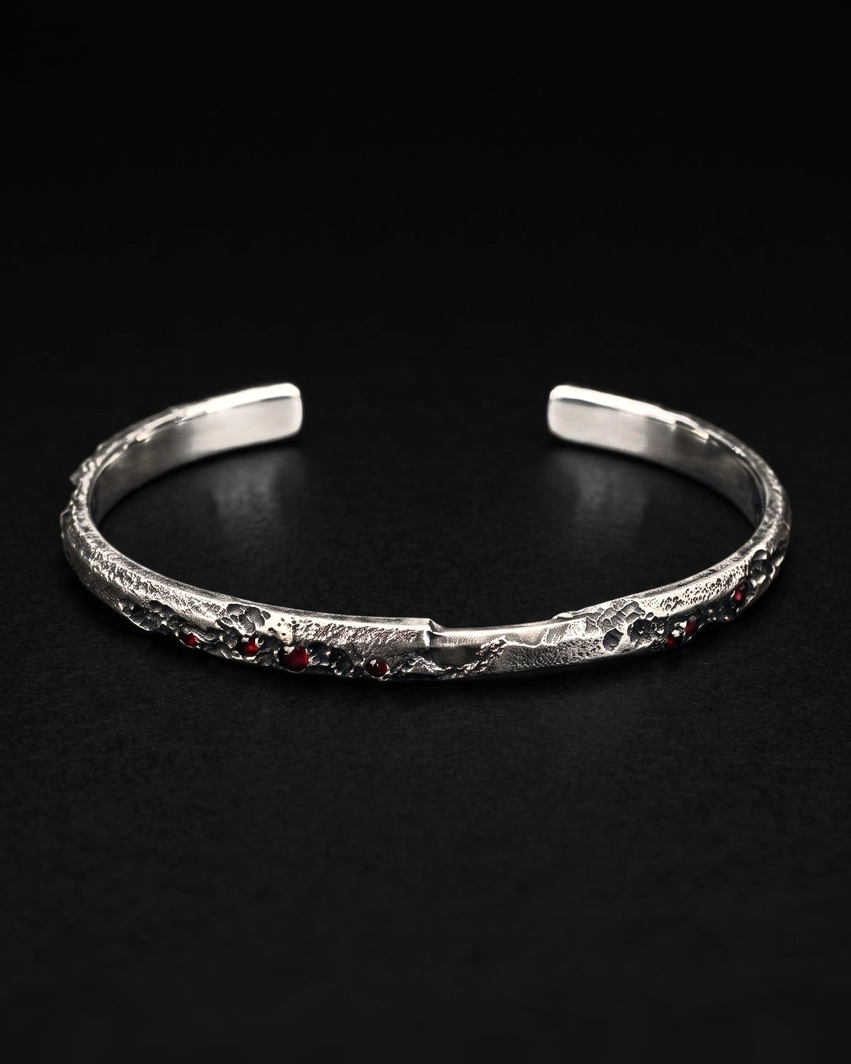 Raw III Bracelet, an open cuff of solid silver with small dark red stones set into its cratered face