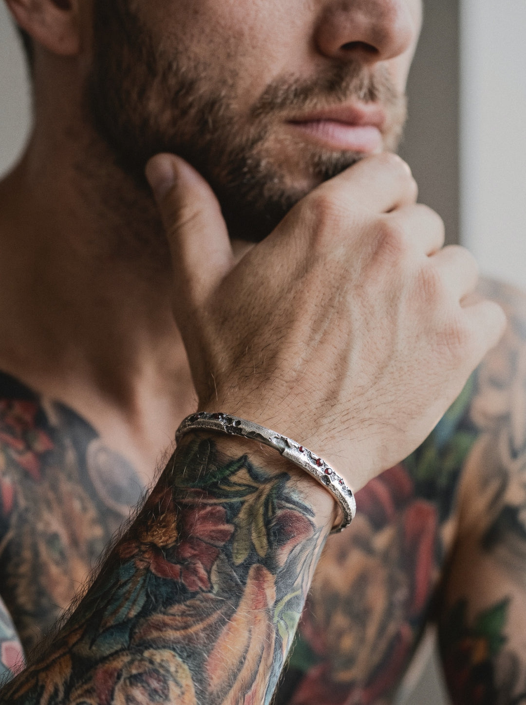 Raw III Bracelet, the cratered silver cuff with small red stones, worn on a man's tattooed wrist, his hand at his beard
