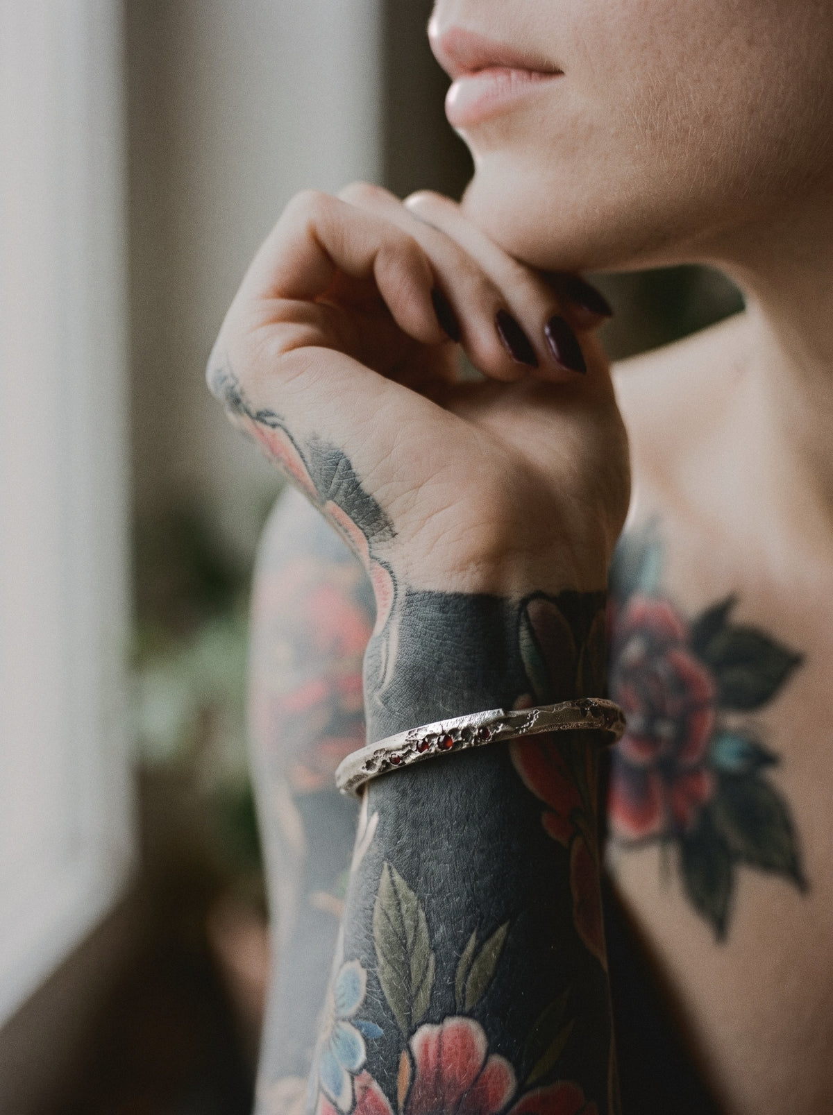 Raw III Bracelet, the cratered silver cuff with small red stones, worn on a woman's tattooed wrist by a window