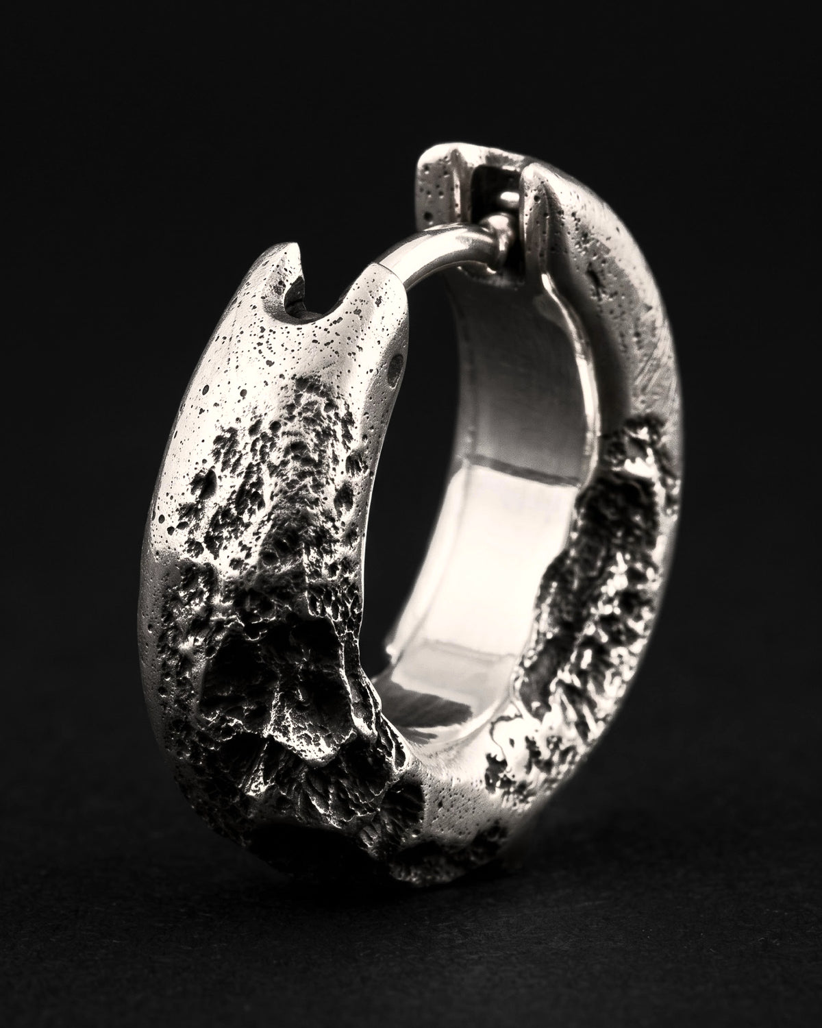 Raw III Earring, a thick cratered silver huggie hoop standing on its edge, three-quarter view against black