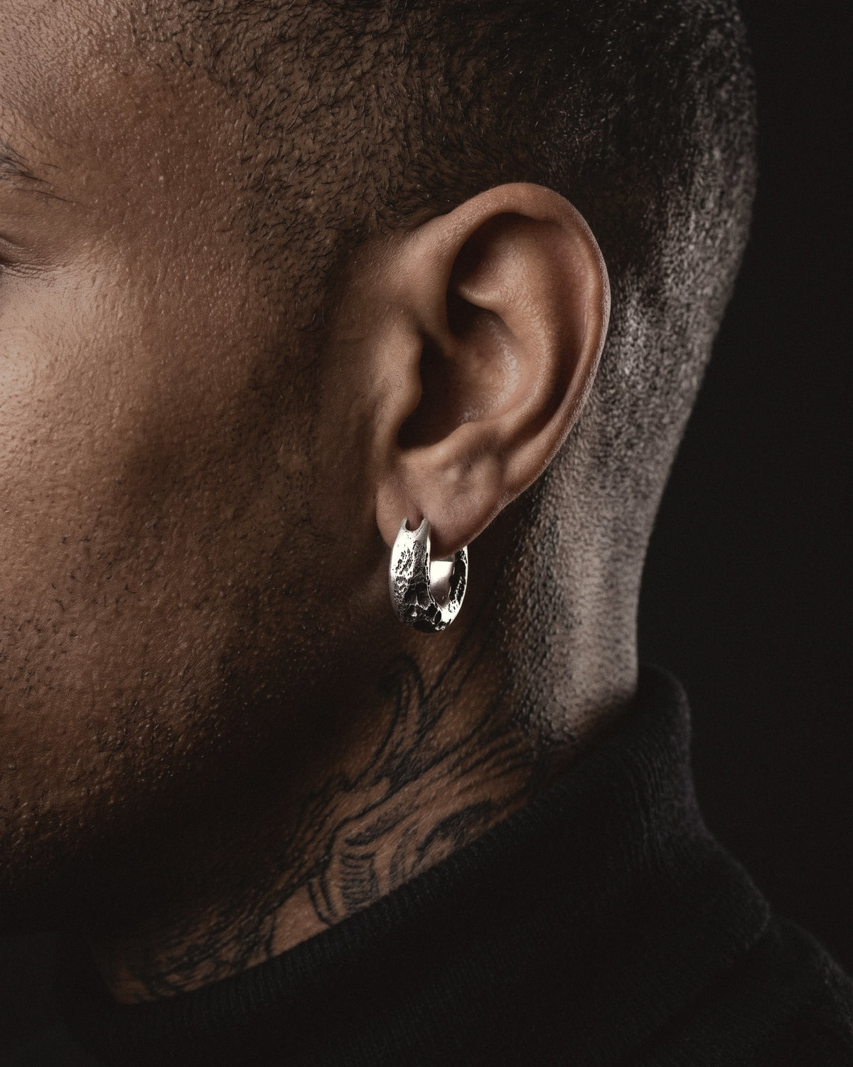 Raw III Earring worn on a man's left ear, a small cratered silver hoop above a tattooed neck and a black turtleneck