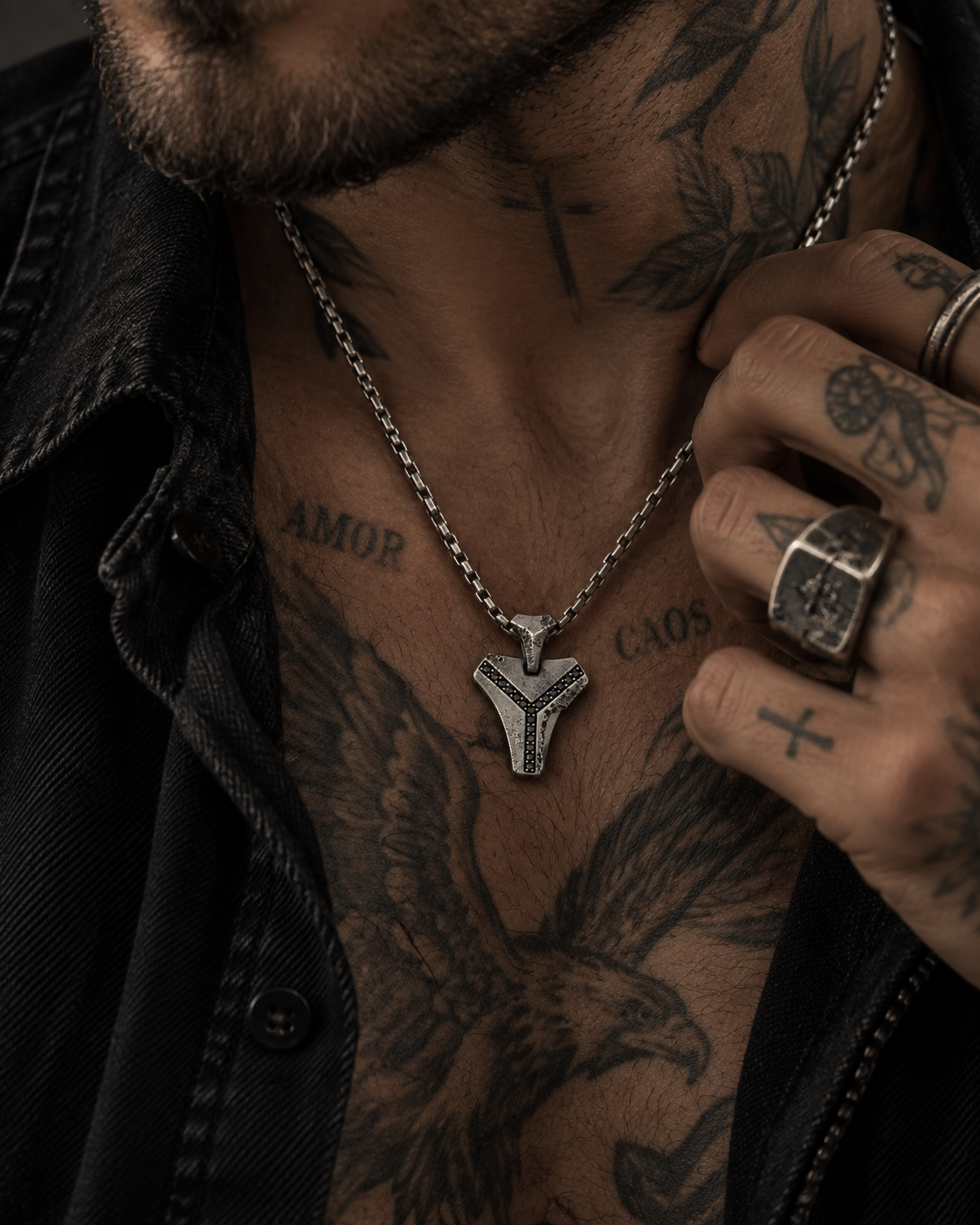 Raw III Pendant worn by a man with a tattooed chest, open black jacket and a ringed hand at the collar