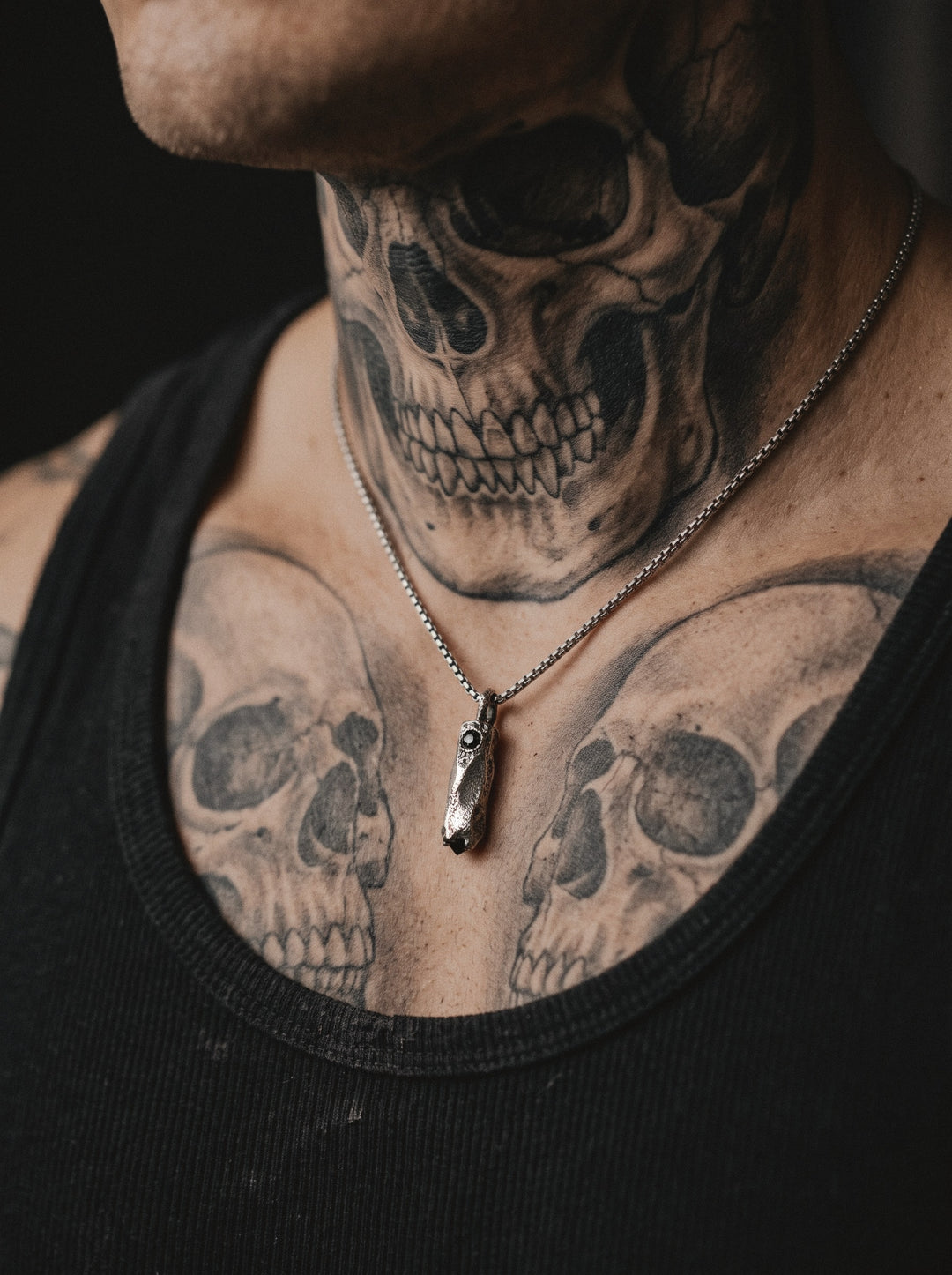Raw V Pendant on a box chain, the small faceted bar worn on a man's chest tattooed with skulls, above a black tank top