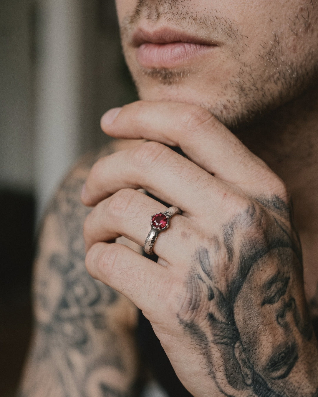 Raw V Ring with its ruby in raw silver prongs, worn on a man's tattooed hand resting at his chin