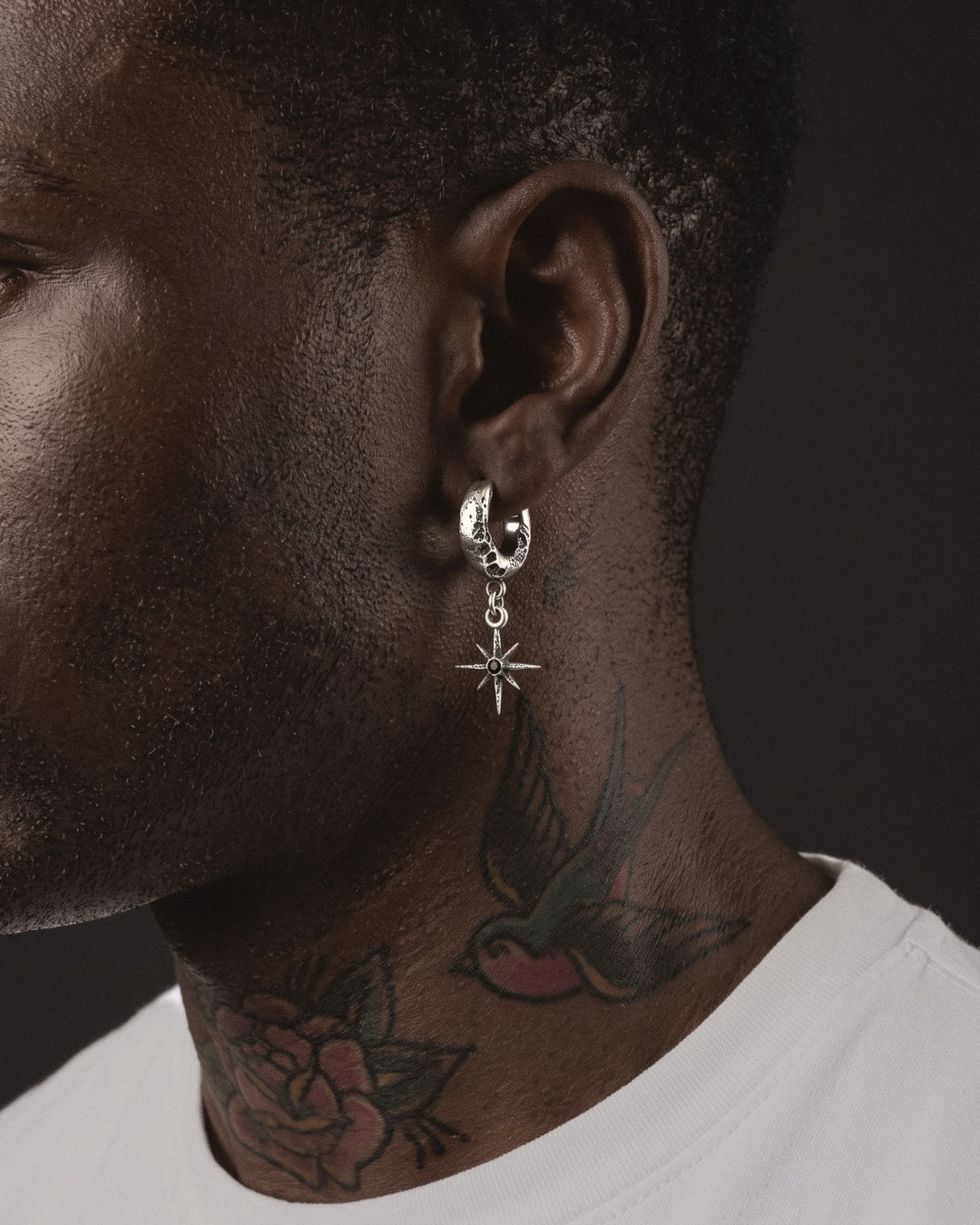 Raw VI Earring worn by a man, the small star with its black stone hanging below the cratered silver hoop, above a swallow tattoo