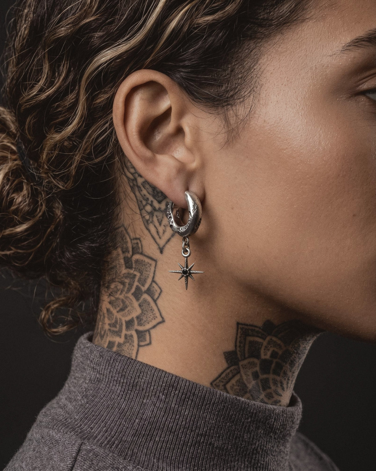 Raw VI Earring worn by a woman, the cratered silver hoop and small star against blackwork mandala tattoos and a grey turtleneck