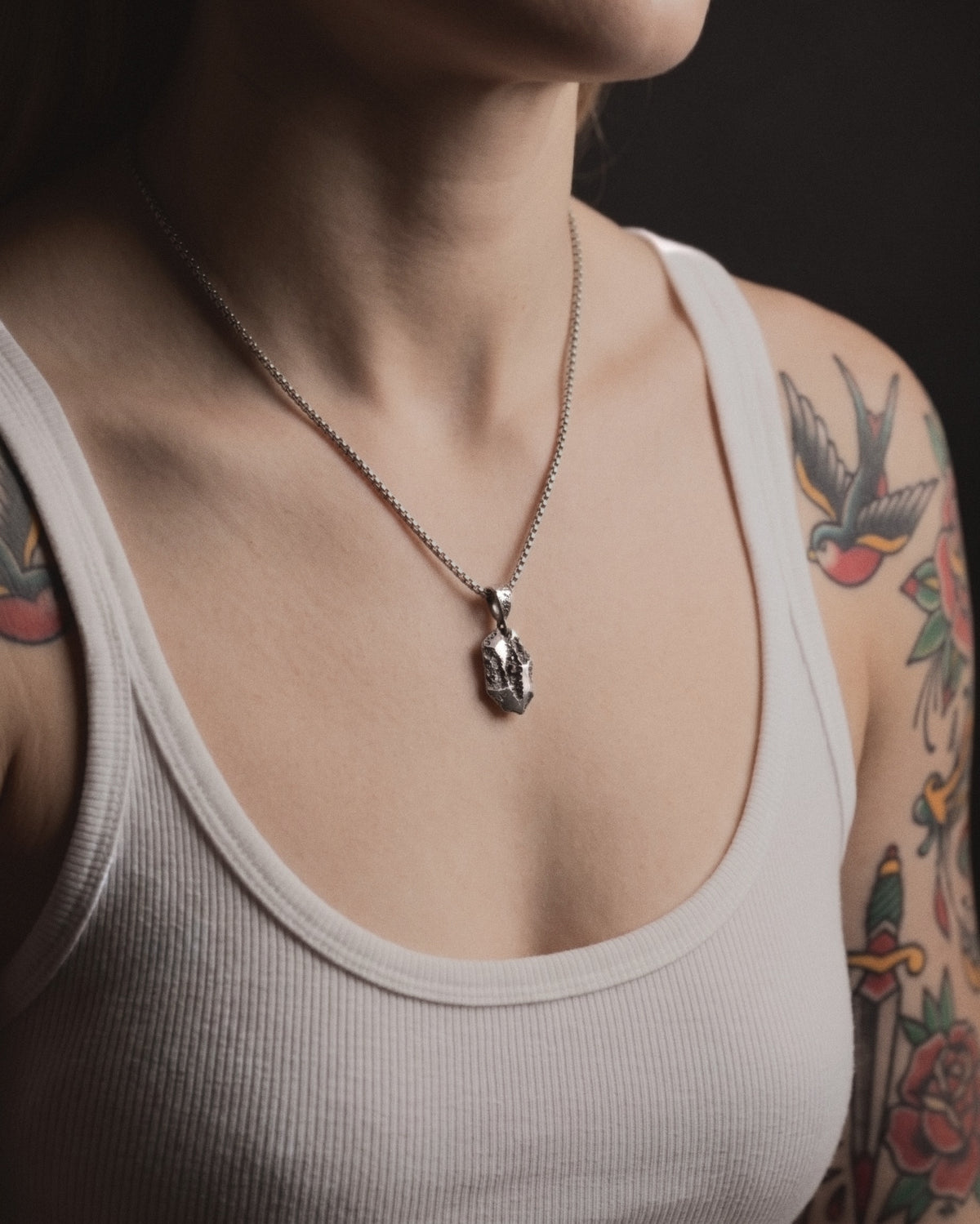 Raw VI Pendant on a box chain, worn on a tattooed woman's chest above a white ribbed tank top