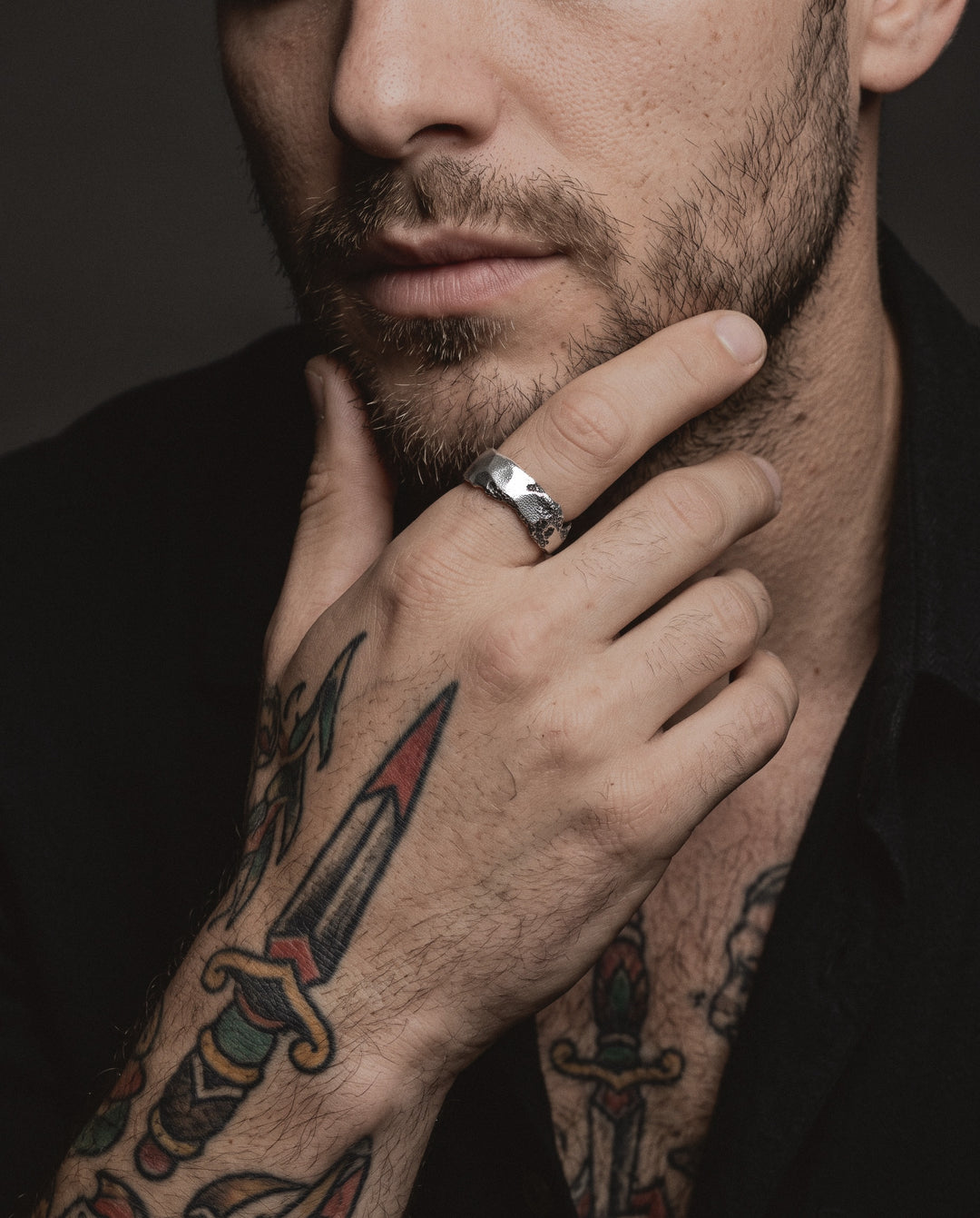 Raw VI Ring worn by a man on his index finger, his hand at his chin, traditional dagger and rose tattoos on his hand, forearm and chest