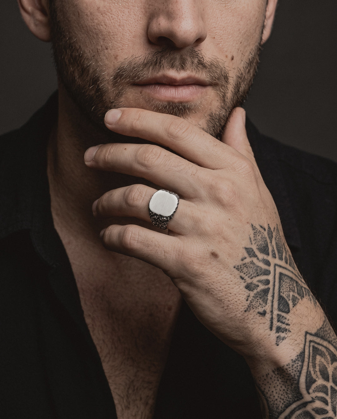 Raw VII Ring worn by a man, his hand at his chin, blackwork and dotwork tattoos on the back of his hand and forearm