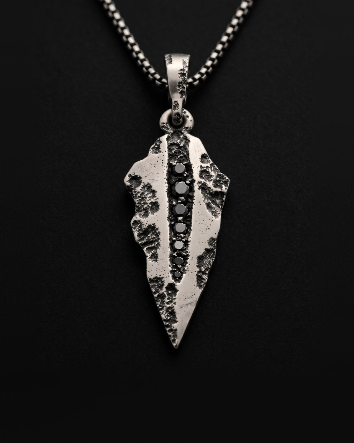 Raw VIII Pendant, a faceted silver arrowhead with seven black stones down a cratered channel, on a box chain against black