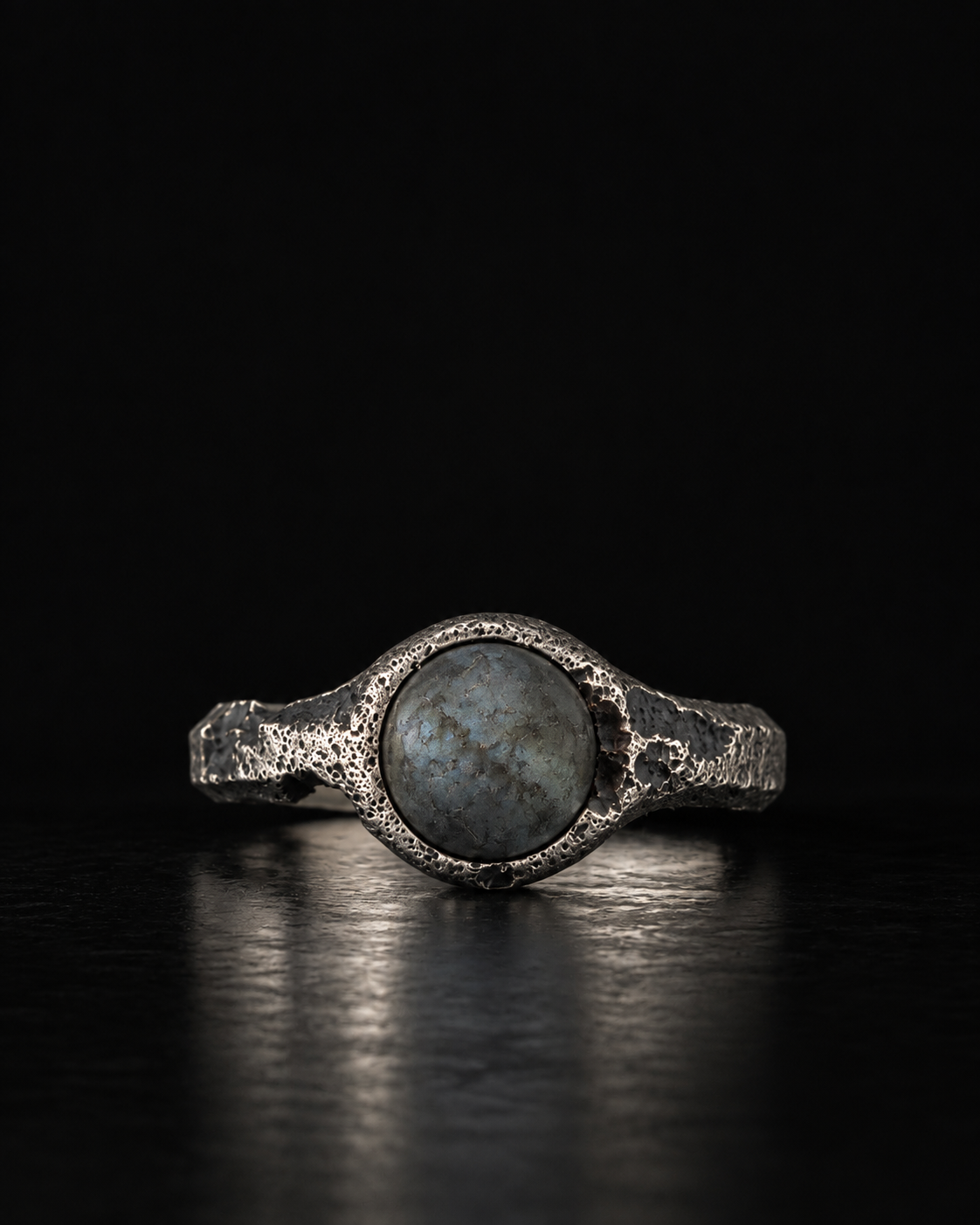 Raw VIII Ring with a round grey labradorite cabochon in a pitted silver bezel, on a black surface