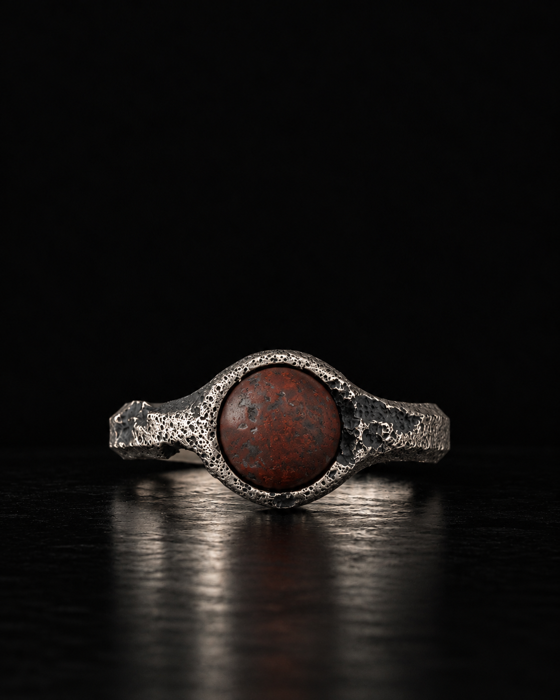 Raw VIII Ring with a round red brecciated stone in a pitted silver bezel, on a black surface