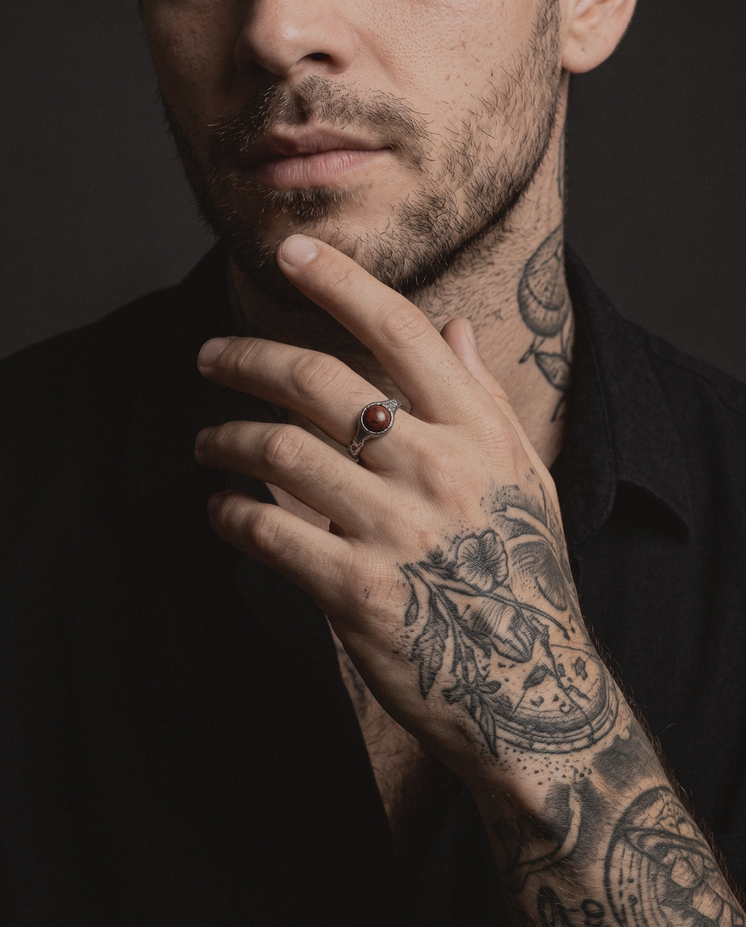 Raw VIII Ring with the red brecciated stone worn by a man, his hand at his chin, etching style tattoos on his hand, arm and neck