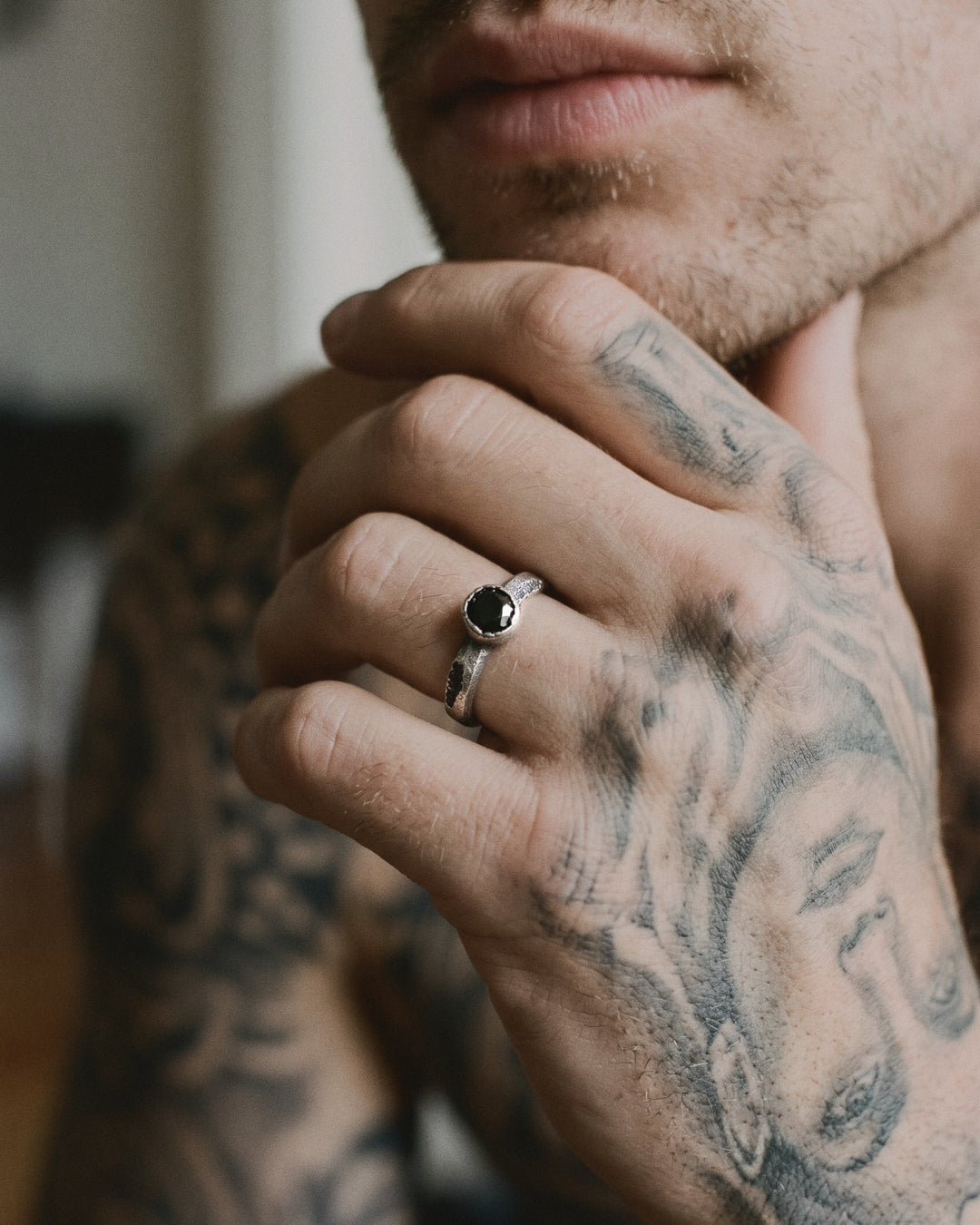 Raw XI Ring with its round black stone, worn on a man's tattooed hand resting at his chin
