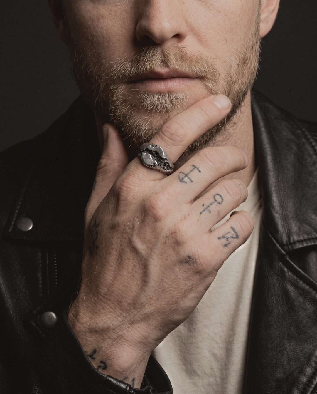 Raw XIV Ring worn on a man's index finger, his hand at his chin, small handpoke tattoos on his fingers