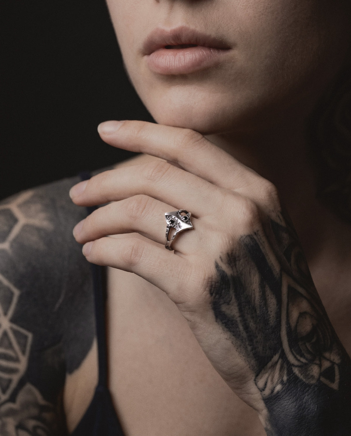 Raw XIX Ring worn by a woman, her hand at her chin, blackwork tattoos on her hand and shoulder