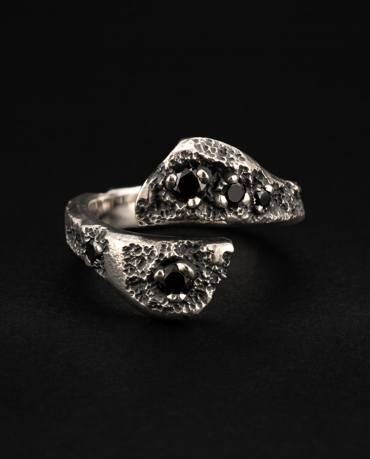Raw XVII Ring face on, an open bypass ring with two flat cratered heads set with black stones, on black
