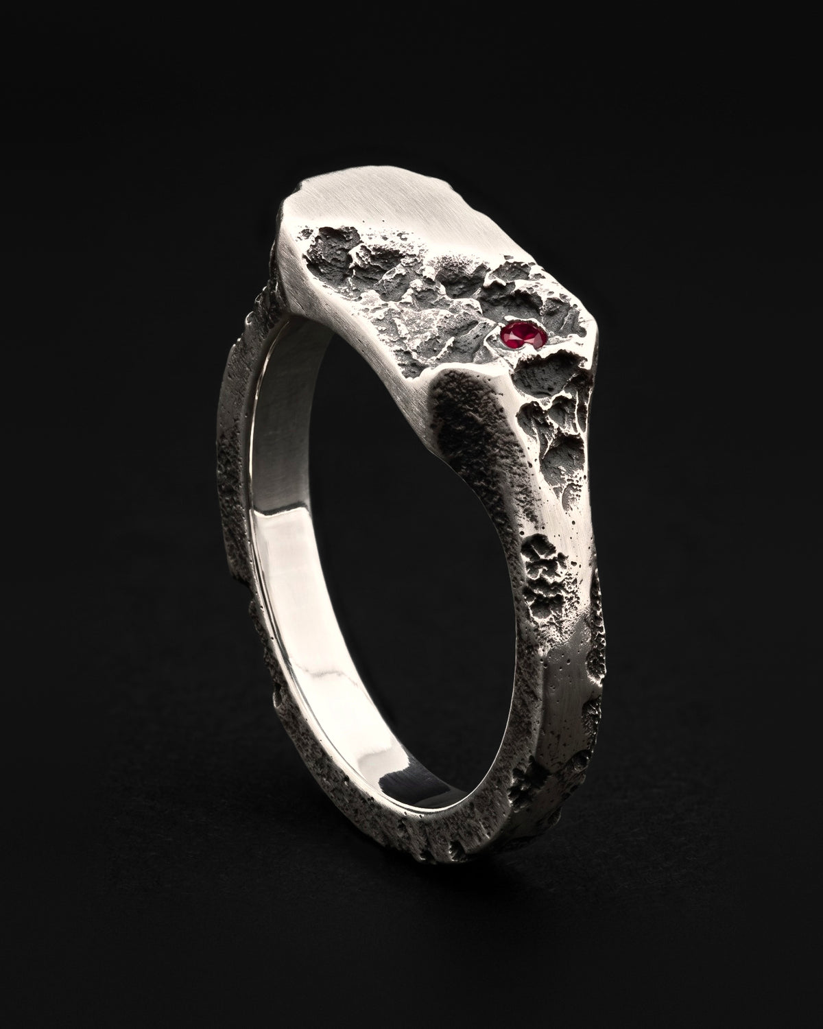 Raw XVIII Ring standing upright at an angle, the bar-shaped face half smooth and half cratered, with a small red stone