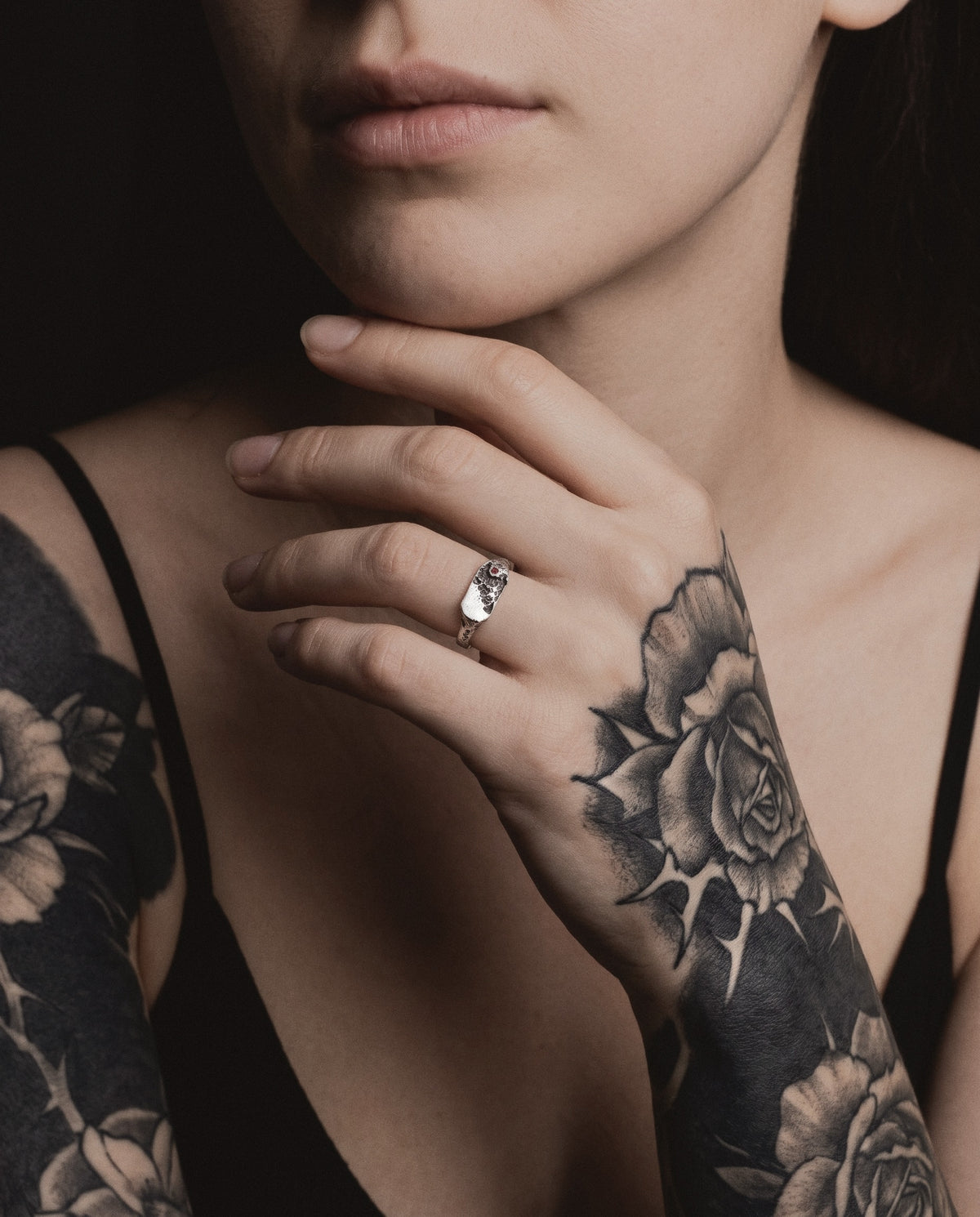 Raw XVIII Ring worn by a woman in black, her hand at her chin, black and grey rose tattoos on her hand and arms