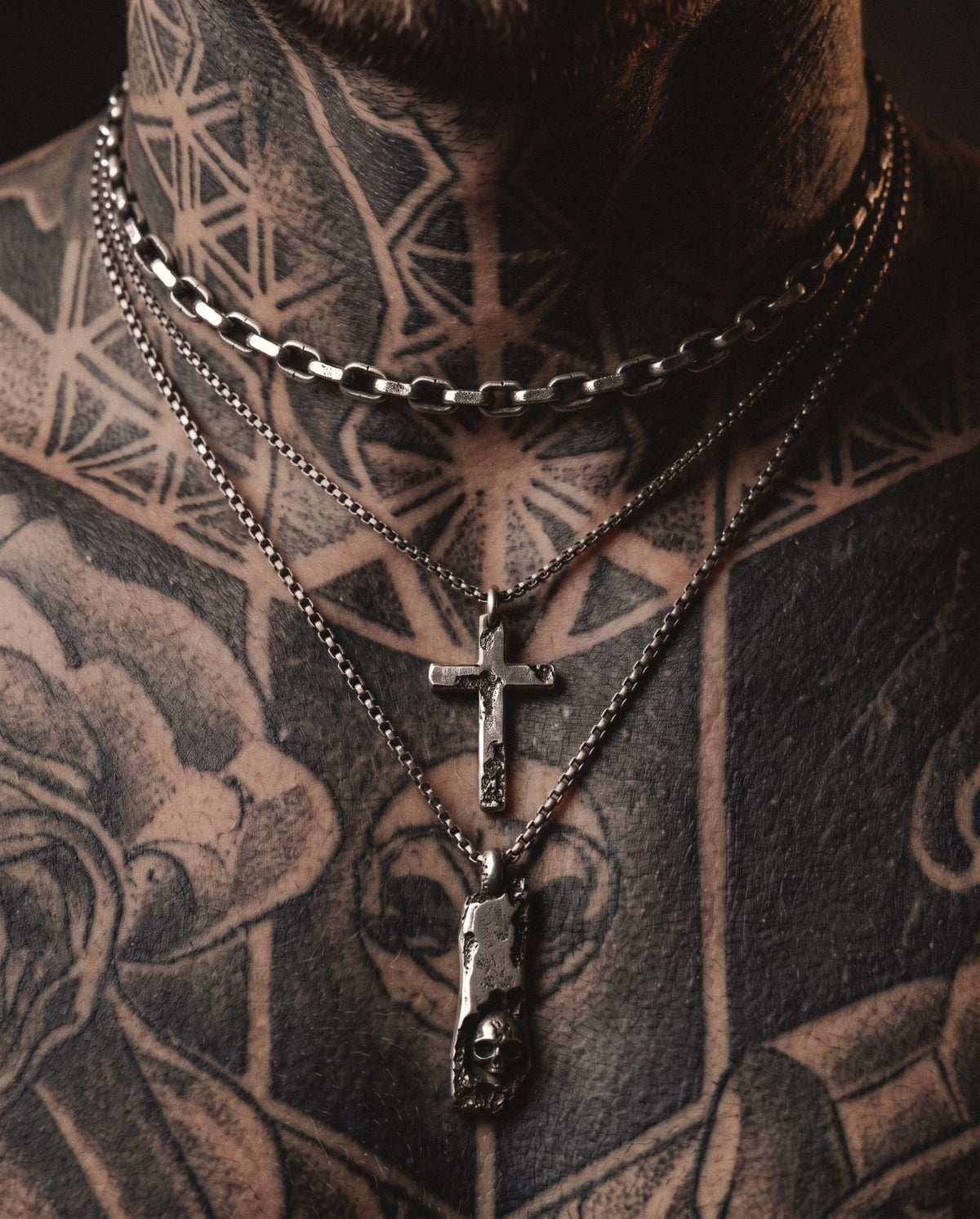 Blackwork tattooed chest wearing a short cratered link necklace, a small cross pendant and a skull bar pendant in raw silver