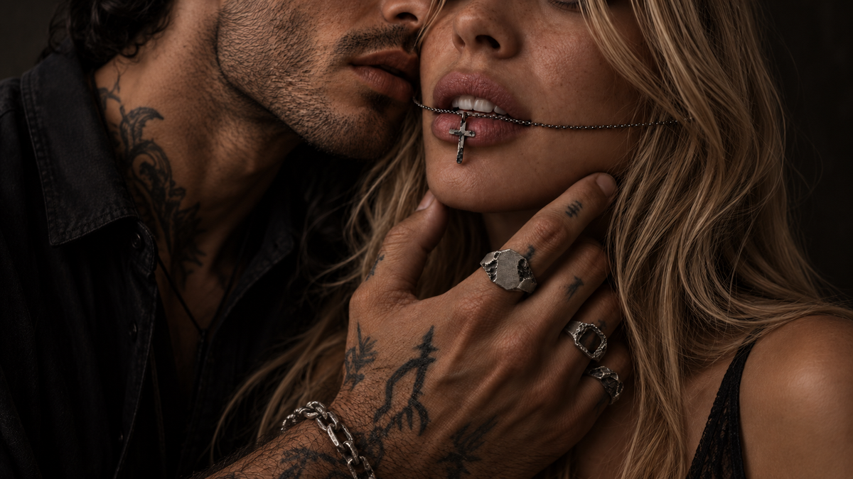 Man and woman close together, his tattooed hand at her jaw wearing three silver rings and a chain bracelet