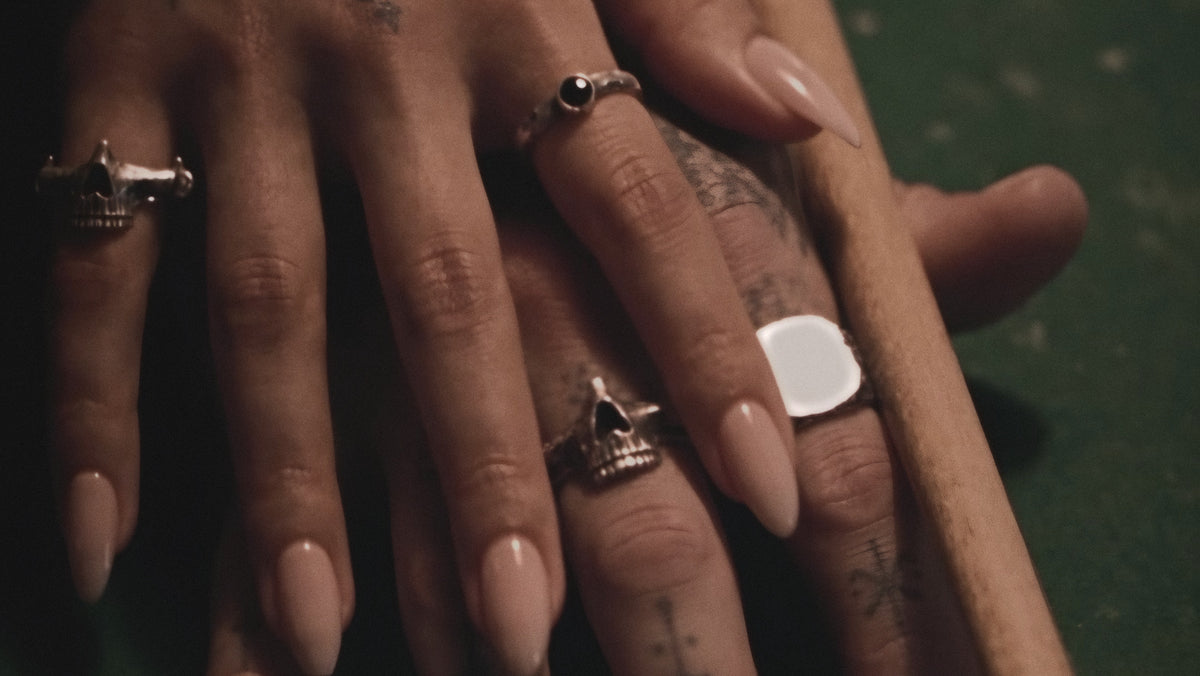 Her hand resting on his on a pool table beside the cue. She wears a skull ring and a thin ring with a black stone; he wears a skull ring and a flat silver signet.