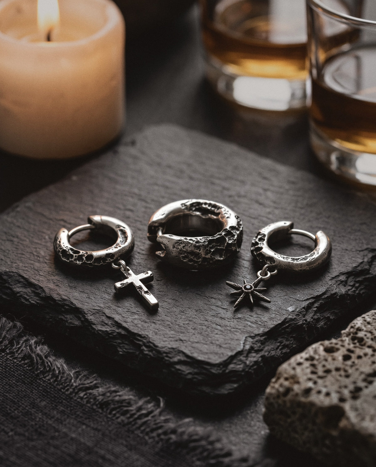 Three cratered silver hoop earrings, one with a cross and one with a star, on dark slate by candlelight
