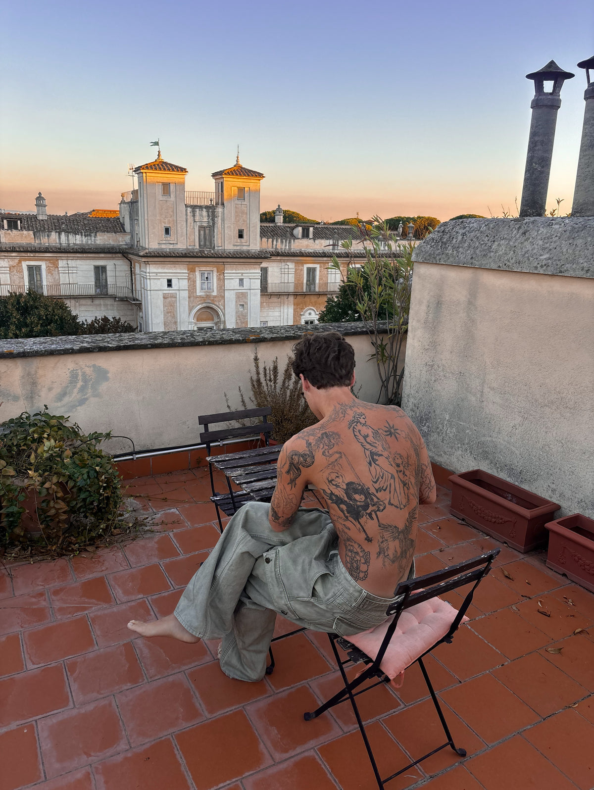 Man with a tattooed back sitting shirtless on a rooftop terrace at sunset