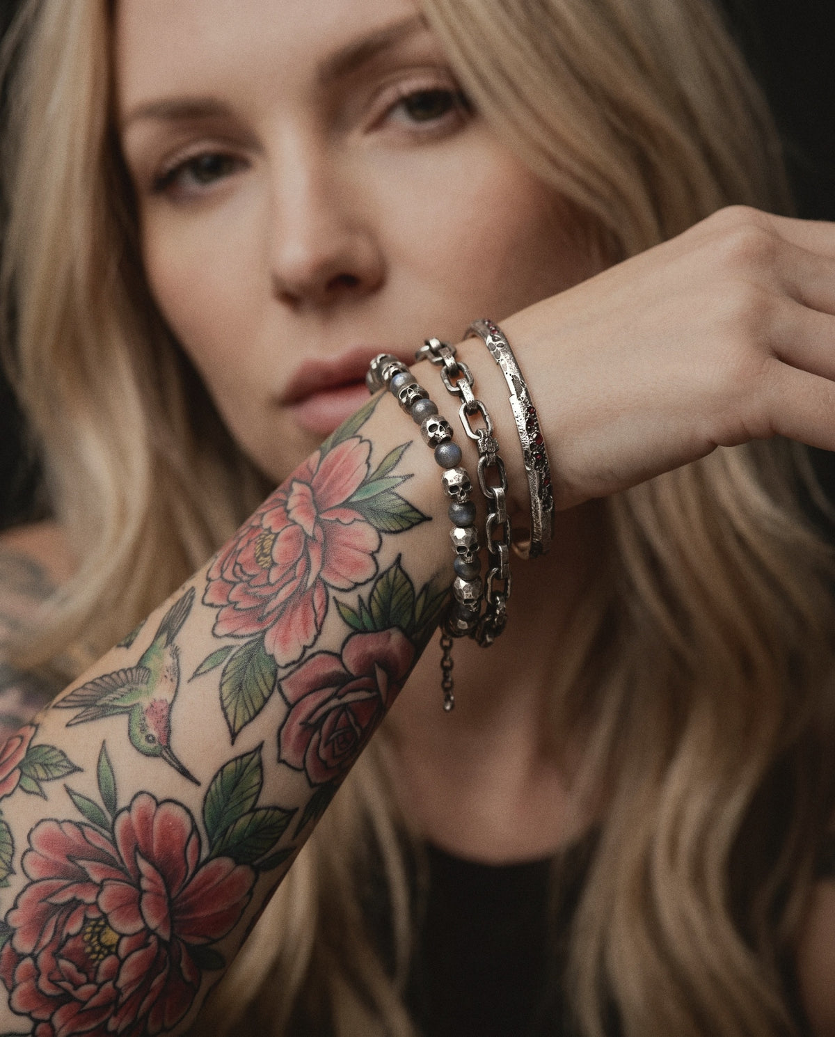 Blonde woman with a colour floral tattoo sleeve wearing a skull and labradorite bead bracelet, a cratered link chain and a ruby cuff in raw silver