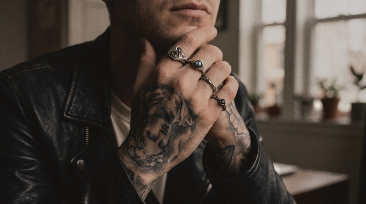 Tattooed hand at the jaw wearing a ruby signet, a labradorite ring, a cratered band and a black spinel ring in raw silver