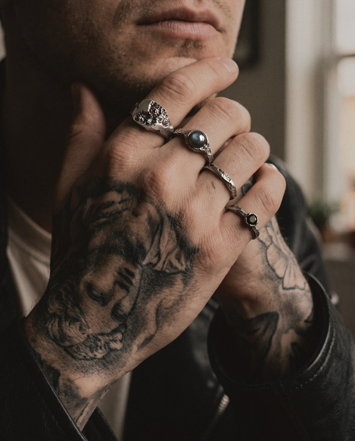 Tattooed hand at the jaw wearing a ruby signet, a labradorite ring, a cratered band and a black spinel ring in raw silver