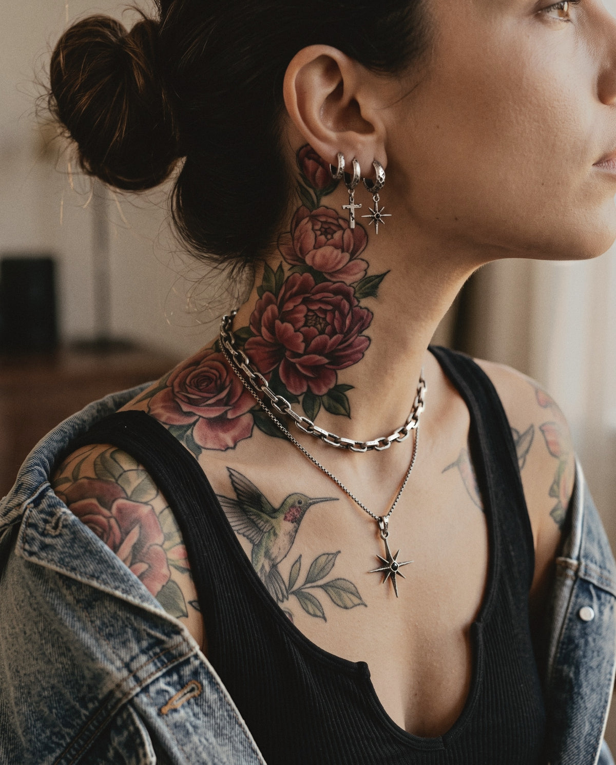 Close-up of a tattooed neck with three cratered silver hoop earrings, a small cross and a star with a black stone, above a heavy link necklace and a star pendant