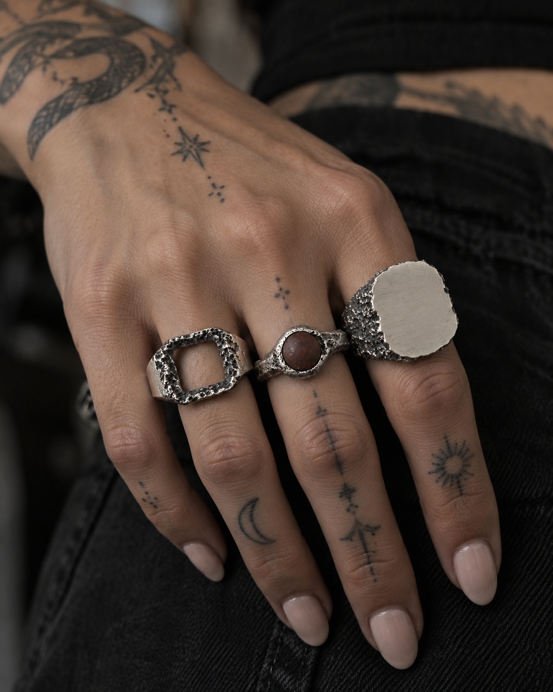 Tattooed woman's hand wearing an open square ring, a brown stone ring and a plain signet